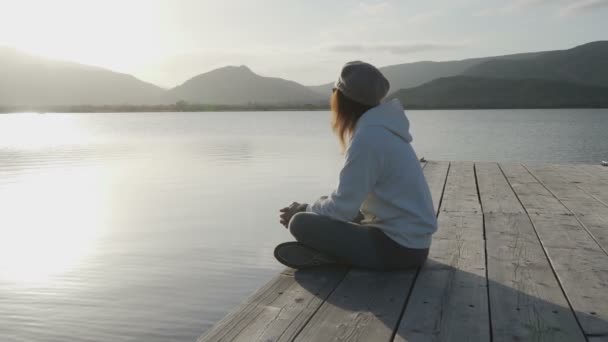 脇から見た長い髪の若い女性は、古い木製の桟橋に座って日没時に湖を見ている - 映像、動画