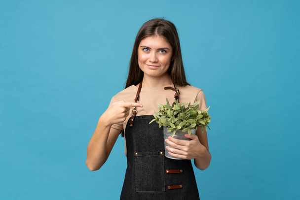 Jeune femme sur fond bleu isolé prenant un pot de fleurs et le pointant
 - Photo, image