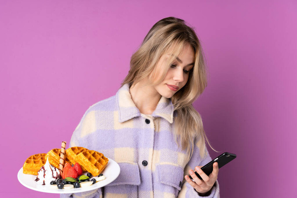 Teenager Russian girl holding waffles isolated on purple background holding coffee to take away and a mobile - Photo, Image