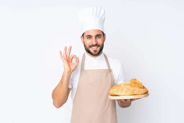 Boulanger masculin tenant une table avec plusieurs pains isolés sur fond blanc montrant un signe ok avec des doigts - Photo, image