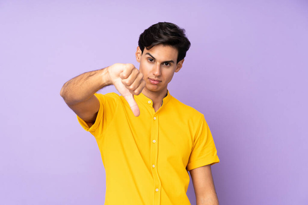 Mężczyzna na pojedynczym fioletowym tle pokazuje kciuk w dół z negatywnym wyrazem - Zdjęcie, obraz
