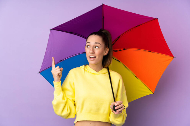 Nuori brunette nainen pitämällä sateenvarjo yli eristetty violetti seinä aikoo toteuttaa ratkaisun nostaessaan sormea ylös
 - Valokuva, kuva