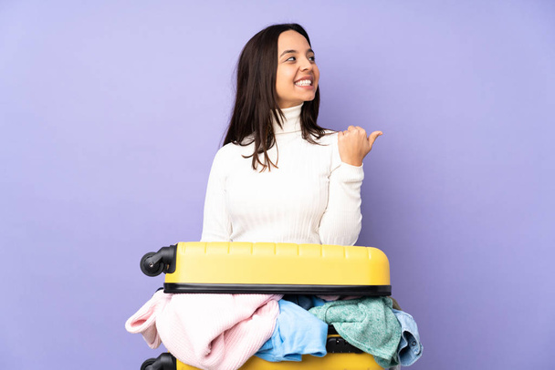 Traveler young woman with a suitcase full of clothes over isolated purple background pointing to the side to present a product - Фото, изображение
