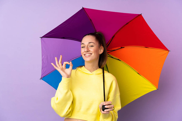 Молодая брюнетка с зонтиком над изолированной фиолетовой стеной показывает знак ОК с пальцами
 - Фото, изображение