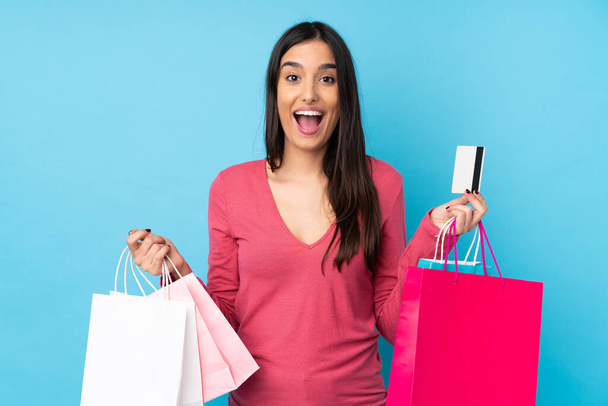 Junge brünette Frau über isoliertem blauem Hintergrund mit Einkaufstaschen und überrascht - Foto, Bild