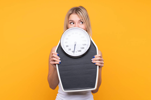 Young blonde Russian woman over isolated yellow background with weighing machine and hiding behind it - Photo, Image