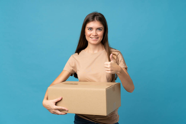 Молода жінка над ізольованим синім тлом тримає коробку, щоб перенести його на інший сайт з великим пальцем вгору
 - Фото, зображення