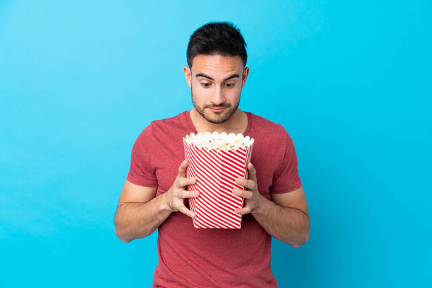 młody przystojny mężczyzna na odizolowanym niebieskim tle trzymając duży wiadro popcornów - Zdjęcie, obraz