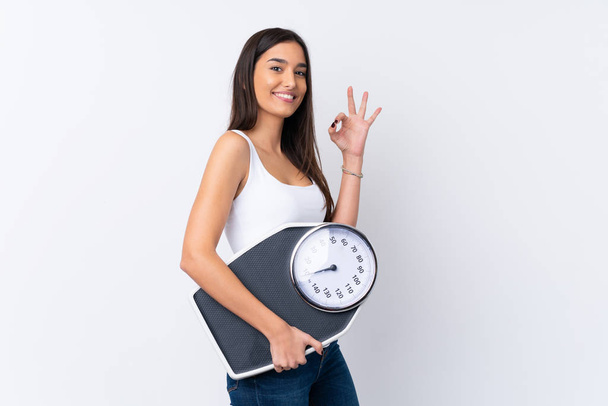 Young brunette woman over isolated white background holding a weighing machine and doing OK sign - Foto, Imagen