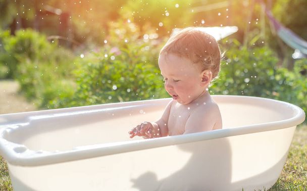 Little baby washes in the bathroom on the green grass. Little baby having fun, splash water and laughs. Happy child bathing outdoors on green grass in spring garden. Summer, vacation, health. - Photo, Image