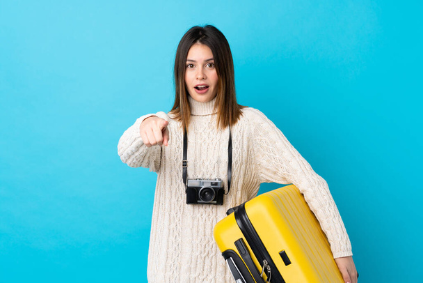 Странница, держащая чемодан над изолированной голубой стеной удивлена и указывает вперед
 - Фото, изображение