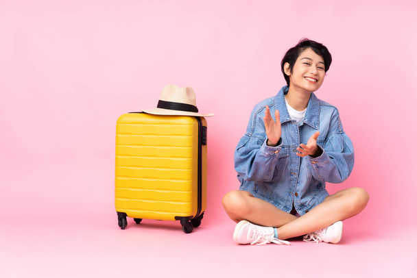 Young traveler Vietnamese woman with suitcase sitting on the floor over isolated pink background applauding after presentation in a conference - 写真・画像