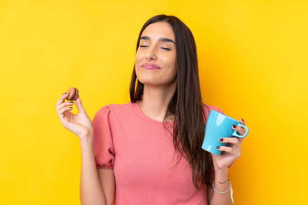 Young brunette woman over isolated yellow background holding colorful French macarons and a cup of milk - Photo, Image