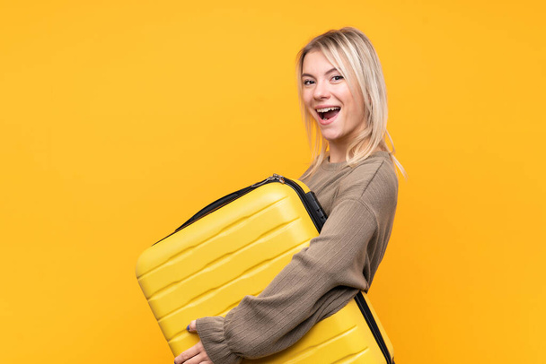 Młoda blondynka na odizolowanym żółtym tle na wakacjach z walizką podróżną - Zdjęcie, obraz