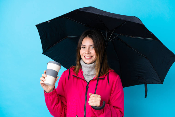 Молодая женщина держит зонтик и кофе, чтобы увезти за изолированную голубую стену
 - Фото, изображение