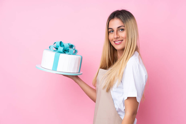 Young Uruguayan pastry woman with a big cake over isolated pink background smiling a lot - Photo, Image