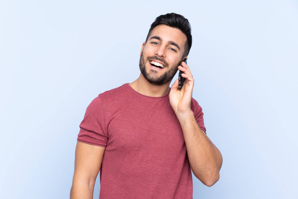 Молодий красивий чоловік з бородою над ізольованим синім тлом, що веде розмову з мобільним телефоном
 - Фото, зображення
