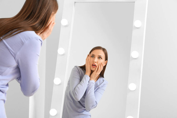 Δυσαρεστημένη νεαρή γυναίκα κοιτάζεται στον καθρέφτη στο σπίτι - Φωτογραφία, εικόνα