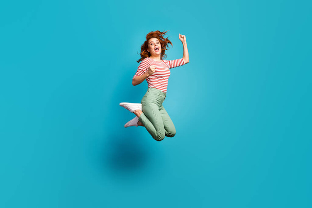 Yeah. Full body profile photo of funny lady jump high feel crazy emotions raise fists win competition wear casual red white shirt green pants footwear isolated blue color background - Foto, imagen