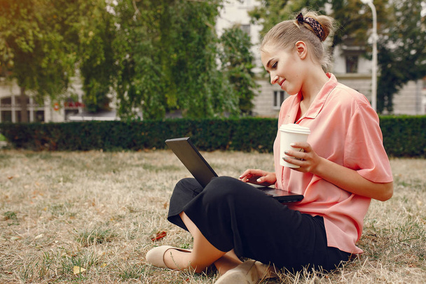 Pembe tişörtlü kız parkta oturuyor ve bilgisayarı kullanıyor. - Fotoğraf, Görsel
