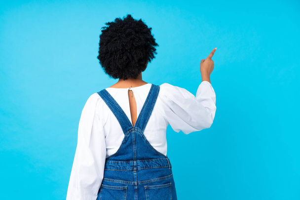 Afrikai amerikai nő overállal elszigetelt kék háttér mutatva vissza a mutatóujját - Fotó, kép