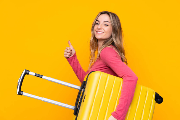 Молодая женщина на изолированном желтом фоне в отпуске с чемоданом и с большим пальцем вверх
 - Фото, изображение