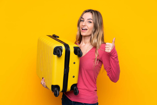 Young woman over isolated yellow background in vacation with travel suitcase and with thumb up - Photo, Image