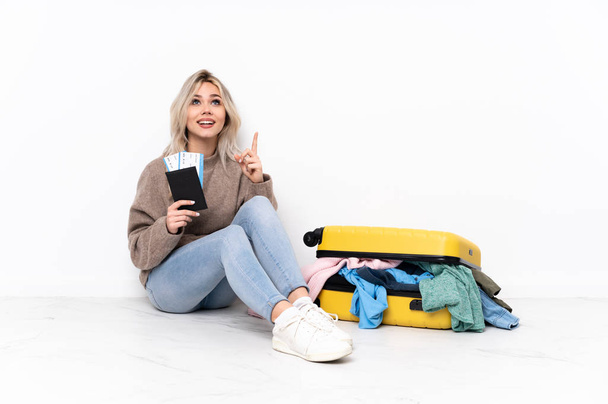 Подростковая блондинка с чемоданом, полным одежды, сидящей на полу, указывая вверх и удивляясь
 - Фото, изображение