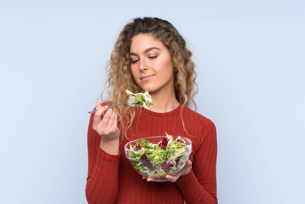 Fiatal szőke nő göndör hajjal salátát tart az elszigetelt falon - Fotó, kép