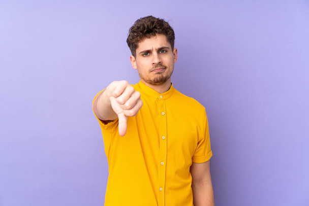 Белый мужчина, выделенный на фиолетовом фоне, показывает большой палец вниз с отрицательным выражением
 - Фото, изображение