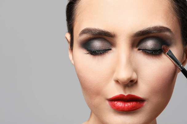 Beautiful young woman applying makeup against grey background - Photo, Image