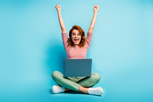 Full size photo of excited ginger hair woman sit floor legs crossed work laptop finish start-up report scream yeah raise fists wear green white sweater footwear isolated blue color background - Foto, afbeelding