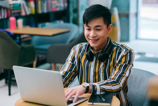 Mirthful young man sitting at the library table and smiling while using a modern laptop - 写真・画像