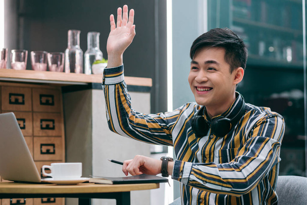Cheerful young man in a cafe putting one hand up while waving to someone in the distance - Foto, Imagem