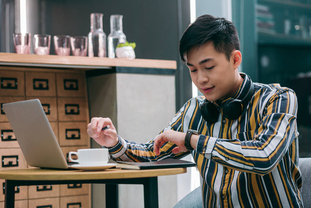 Young man sitting with a laptop in cafe and looking at his watch attentively - 写真・画像