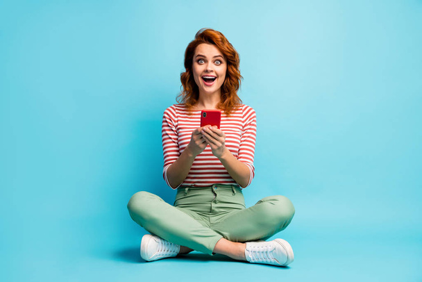 Full size photo of enthusiastic astonished girl sit legs crossed use cellphone read social network news impressed scream wow omg wear green white shoes isolated over blue color background - Foto, Imagem