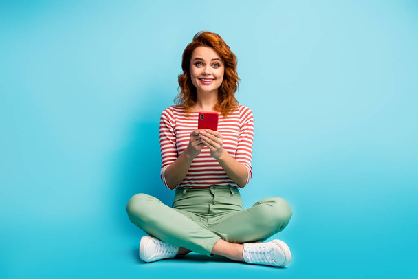 Full length photo of positive cheerful woman sit legs crossed use smartphone follow comment share repost comment social media news wear stylish outfit isolated over blue color background - Photo, image