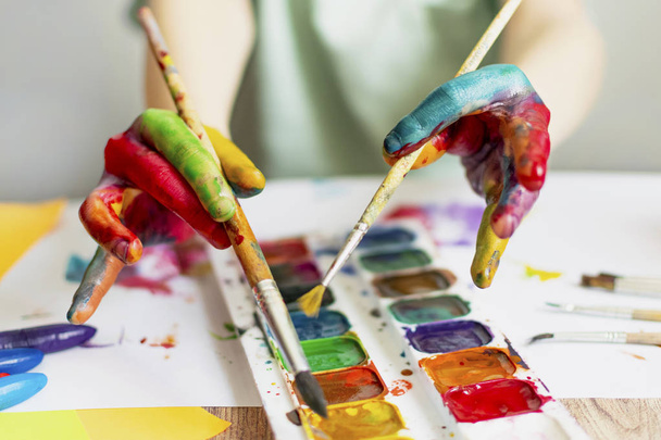 Ребенок рисует. Ладони и руки ребенка в разноцветной акварели. Акварель, гуашь, кисти, цветные и восковые карандаши, наклейки. Набор для рисования, творчества и хобби
 - Фото, изображение