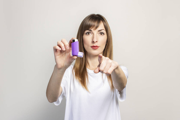 mujer joven sostiene un inhalador en su mano, señala su dedo a la derecha sobre un fondo claro. Banner. Concepto para una respiración más fácil, tratamiento del asma, faringe, laringe, tráquea
. - Foto, Imagen