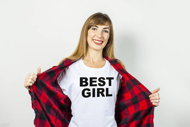 Молодая женщина с улыбкой в красной рубашке и белой футболке на белом фоне. Текст на футболке добавил "Лучшая девушка". Концепция друга, шок, сюрприз. Баннер
. - Фото, изображение