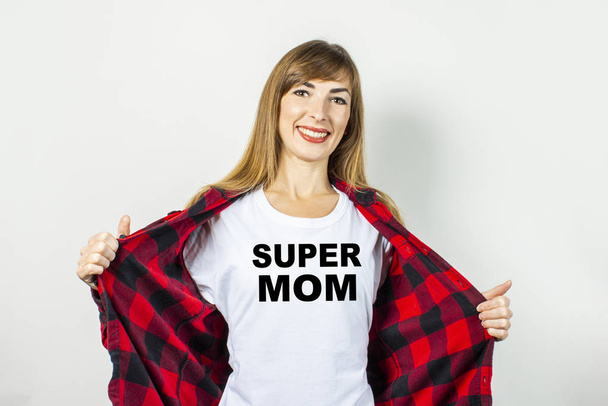 Молодая женщина с улыбкой в красной рубашке и белой футболке на белом фоне. Добавил текст "Супер мама" к рубашке. Концепция текста, логотипа, шока, сюрприза. Баннер
. - Фото, изображение