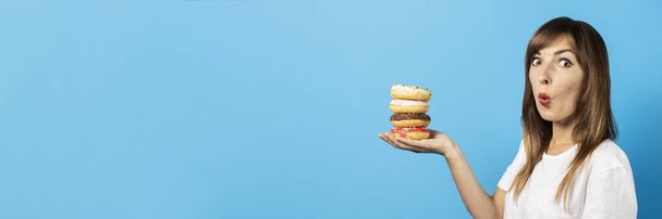 Молодая девушка держит четыре пончика на ладони на синем фоне. Баннер. Концепция диеты, яркие цвета, эмоции. - Фото, изображение