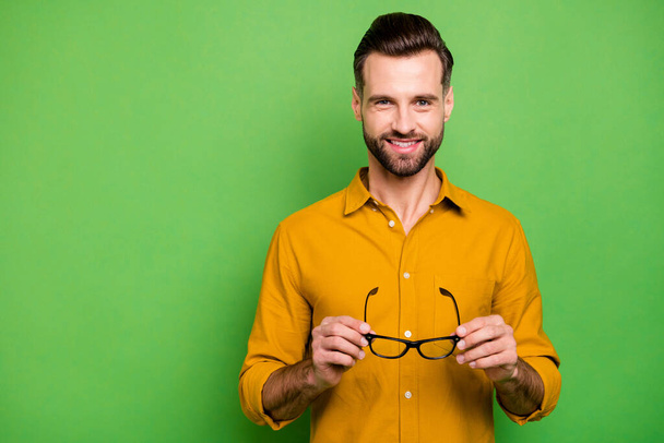 Close-up portrait of his he nice attractive cheerful cheery bearded guy in casual formal shirt holding in hands specs eye care isolated on bright vivid shine vibrant green color background - Foto, Imagem