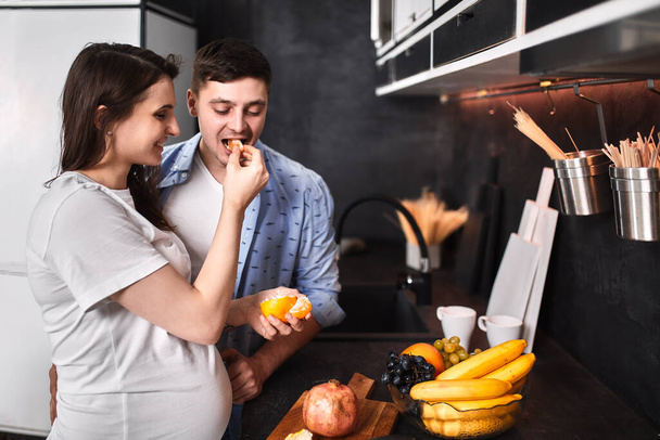 Young pregnant couple in the kitchen, cuddling, preparing food and rejoicing in anticipation of the baby. Concept new life, young family. - Foto, Imagen