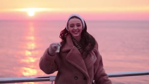 glimlachend jong meisje in bont jas met kopje koffie op de zeepromenade is blij om te zien een mooie zonsopgang - Video