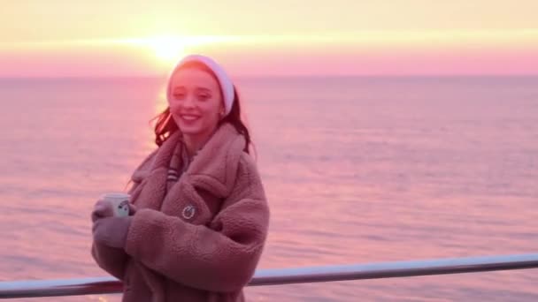 glimlachend jong meisje in bont jas met kopje koffie op de zeepromenade is blij om te zien een mooie zonsopgang - Video