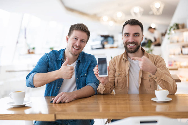 two handsome colleagues men out of office during coffee break at cafe, drinking coffee, pointing at smartphone showing like sign , multitasking concept. - Foto, Imagen