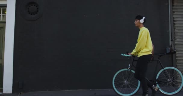Side view of a fashionable mixed race transgender adult hanging out on a summer day in the city, wearing headphones, walking and wheeling his bicycle during a sunny day in slow motion. - Video