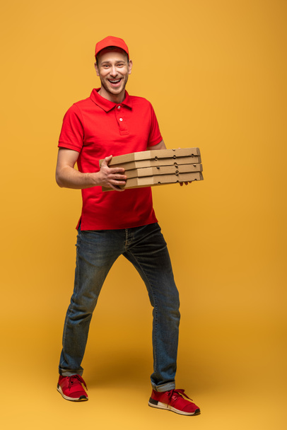 vista completa de hombre entrega feliz en uniforme rojo caminando con cajas de pizza en amarillo
 - Foto, Imagen