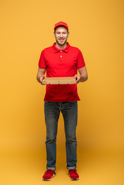 full length view of happy delivery man in red uniform holding pizza box on yellow - Fotoğraf, Görsel